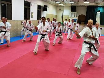 קבוצת מתאמנים בחגורות שחורות מבצעת יחד עמידת קראטה על מזרנים אדומים באולם האימונים